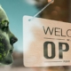 woman's face with technology code behind her facing a "welcome, we're open" sign hanging in a store front window