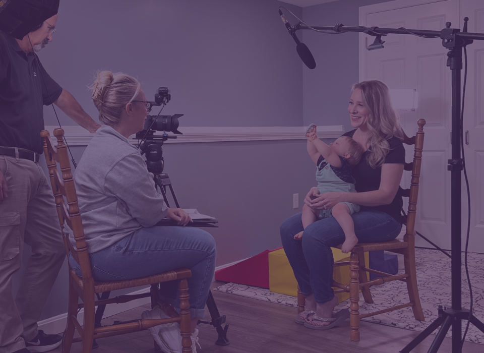 A woman sitting with a baby on her lap as she is interviewed with a camera, microphone and lights all around