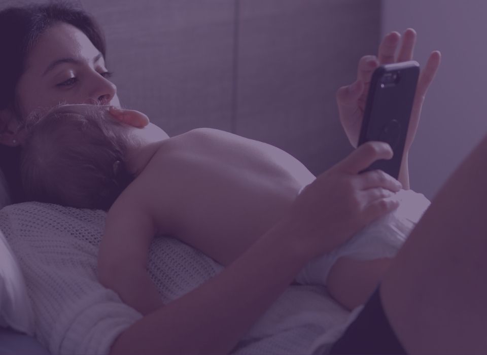 a woman lying back in bed looking at her phone while a baby sleeps on her chest