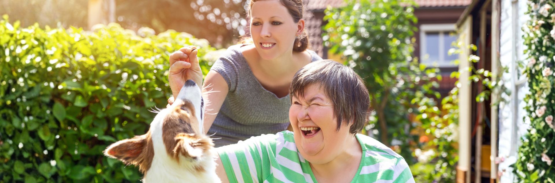 a developmentally disabled woman laughs as she feeds a treat to a jumping dog, with another smiling woman behind her
