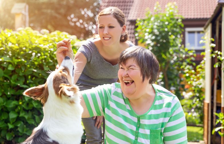 two women laugh as they hold up a treat for a jumping dog